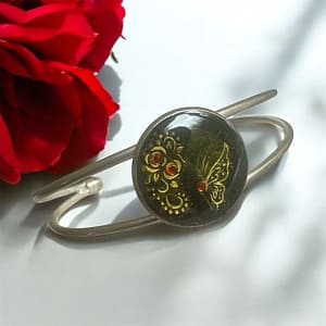 Bracelet jonc en acier avec cabochon en résine noire, décoré de deux roses et d’un papillon orné de strass rouges.